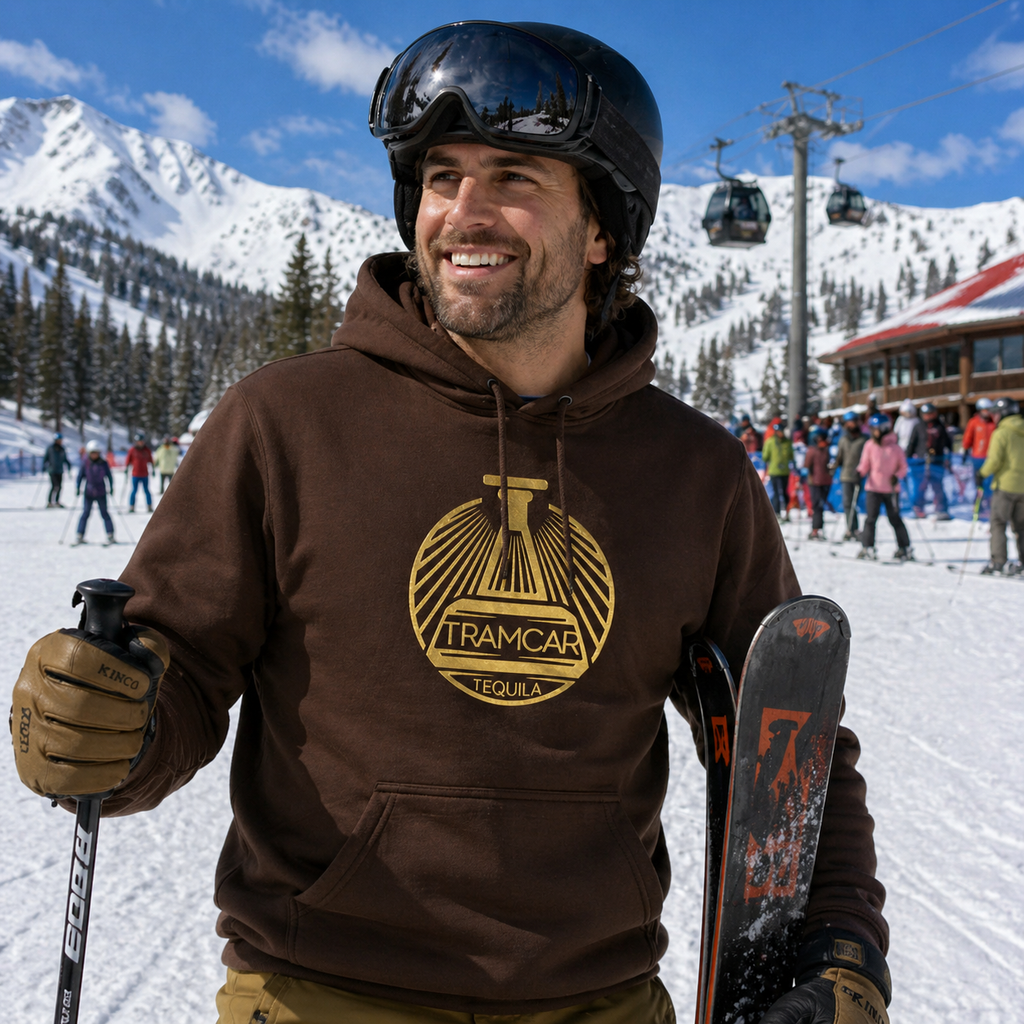 Man wearing a brown Tramcar hoodie holding a snowboard at a ski resort